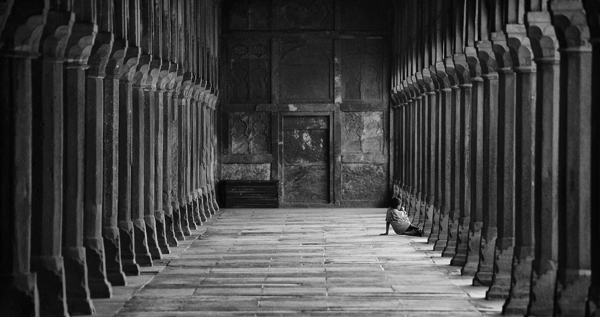 A solitary visitor seated at the far end of a long colonnade of carved stone pillars, light falling across the worn floor.