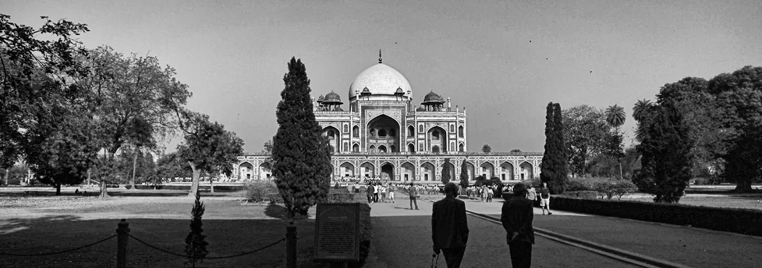Two visitors walking the garden path toward Humayun’s Tomb, its white marble dome rising over the red sandstone facade.
