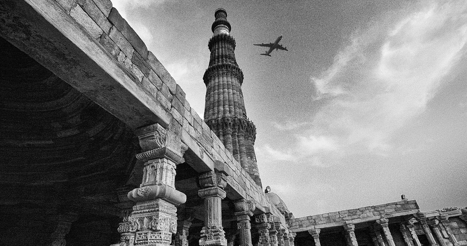 Qutub Minar seen from below through a ruined colonnade, a passenger jet crossing the sky behind it.