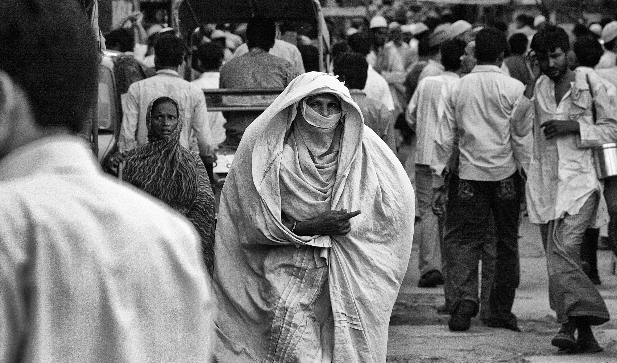 A woman wrapped in white robes moving through the crowd of Nizamuddin Basti, her gaze meeting the camera.